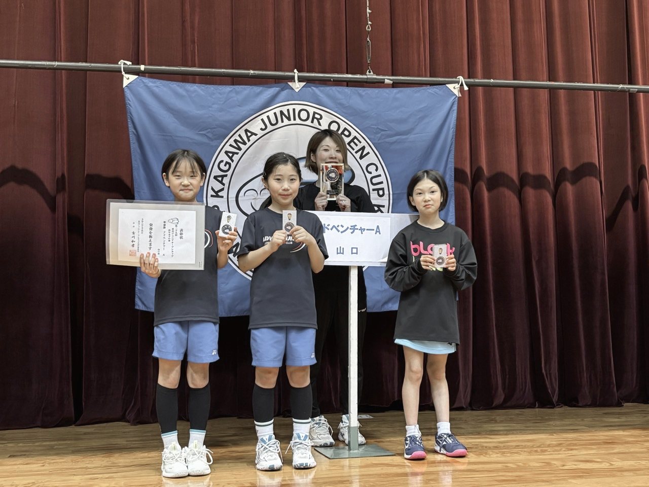 大会の旗の前で、賞状とメダルを持つ子どもたち